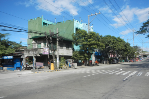 Eine Stadtstraße mit einem grünen Gebäude, fahrenden Fahrzeugen, Fußgängern auf dem Gehweg, Stromleitungen an Masten und einer bewölkten Himmel.