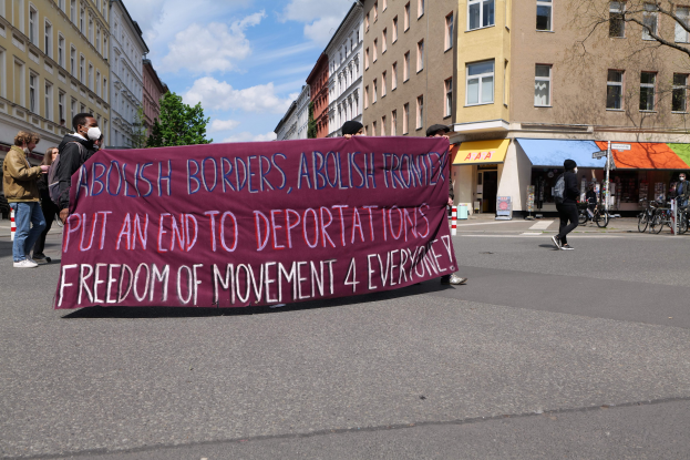 Eine Gruppe von Menschen marschiert mit einem Banner, auf dem "Abolish Borders, Abolish Frontiers, Put an End to Deportations, Freedom of Movement 4 Everyone" steht, durch eine Straße mit Gebäuden, Bäumen, Fahrrádern und bewölktem Himmel im Hintergrund.