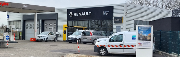 Ein Renault-Autohaus mit parkenden Autos davor, umgeben von einem Metallzaun, einem Schild, Pfählen, einer Texttafel, Bäumen und einem bewölkten Himmel.