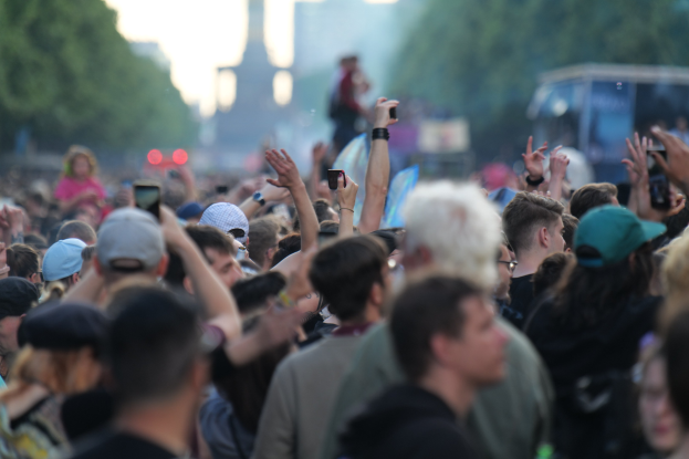 Menschenmenge auf einem Musikfestival mit erhobenen Händen, einige tragen Mützen und halten Handys, vor einem unscharfen Hintergrund aus Bäumen, Gebäuden, Lichtern und einem Fahrzeug.