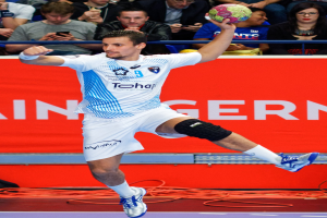 Ein Mann in der Luft mit einem Handball, umgeben von einer Gruppe von Menschen und einem Banner mit Text im Hintergrund.
