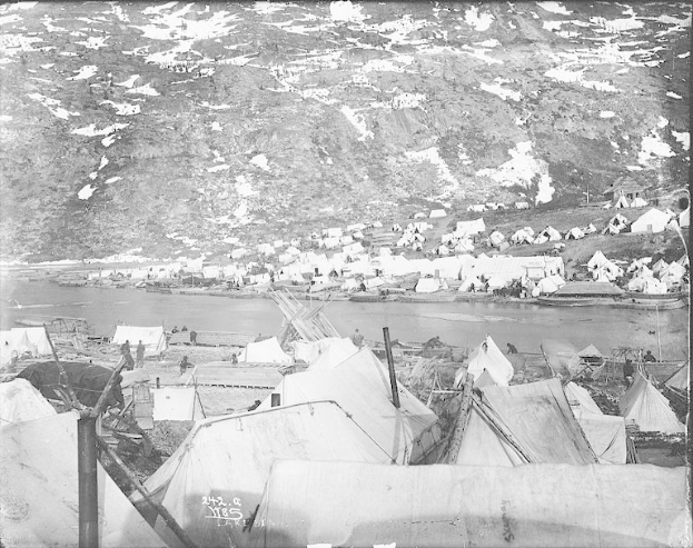 Schwarzes und weißes Foto einer Gruppe von Zelten in einem Camp in der Nähe eines Gewässers mit einem Hügel im Hintergrund.