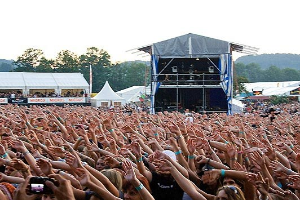 Menge bei einem Musikfestival mit erhobenen Händen und Mobiltelefonen, Bühne im Hintergrund unter Zelten und Bäumen unter einem klaren blauen Himmel.