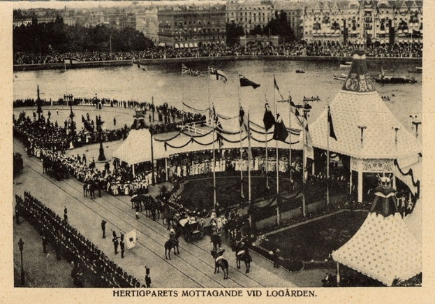 Ein altes Schwarz-Weiß-Foto einer Menge, die sich um ein Faschingszelt mit Fahnen, Stangen und Pferden im Vordergrund und Gebäuden, Bäumen und Wasser im Hintergrund versammelt hat, mit Text am unteren Bildrand.