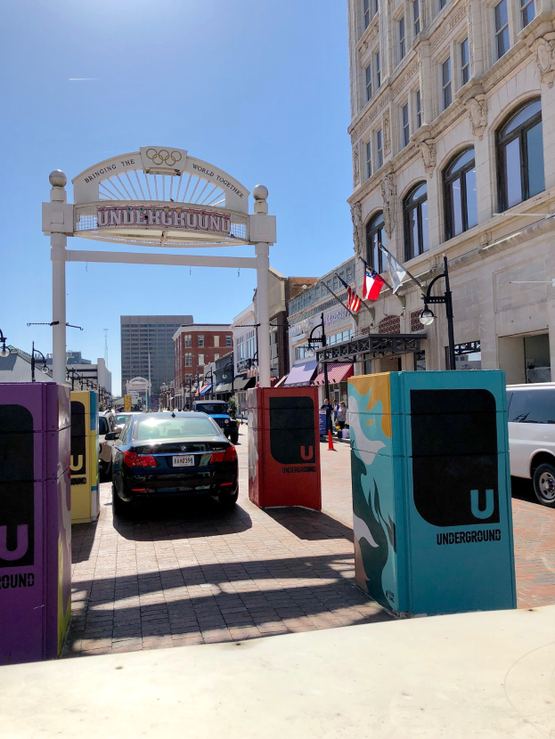 Stadtstraße mit parkenden Autos, einem Torbogen, Gebäuden, Laternen, Fahnenmasten, einem Müllbehälter und Himmel im Hintergrund.