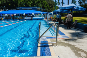 Ein großes Schwimmbad mit schwimmenden Menschen, umgeben von Geländern, Sonnenschirmen, Stühlen, Tischen, Pflanzen, Gras, Bäumen und einem Gebäude im Hintergrund unter einem klaren blauen Himmel.