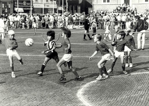 Schwarzes Bild von jungen Jungs, die auf der Straße Fußball spielen, umgeben von einer Zuschauermenge hinter einem Geländer, Gebäuden und Texttafeln im Hintergrund.