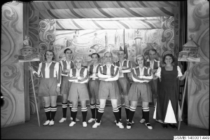 Eine Gruppe lachender Frauen in einem Fußballteam der 1940er Jahre posiert auf einer Bühne, jede hält einen Stock in der Hand, mit einem Bild im Hintergrund.