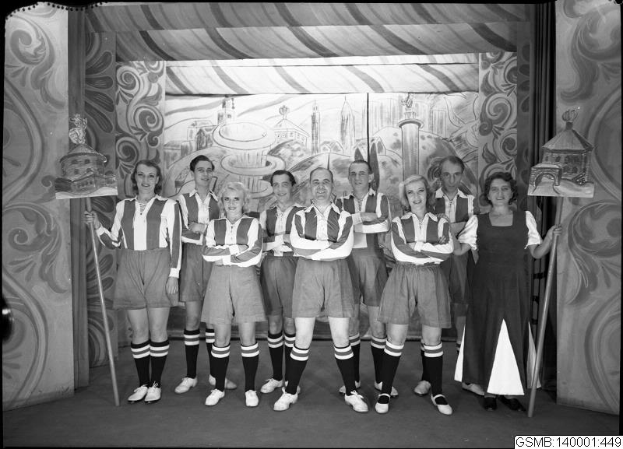 Eine Gruppe lachender Frauen in einem Fußballteam der 1940er Jahre posiert auf einer Bühne, jede hält einen Stock in der Hand, mit einem Bild im Hintergrund.