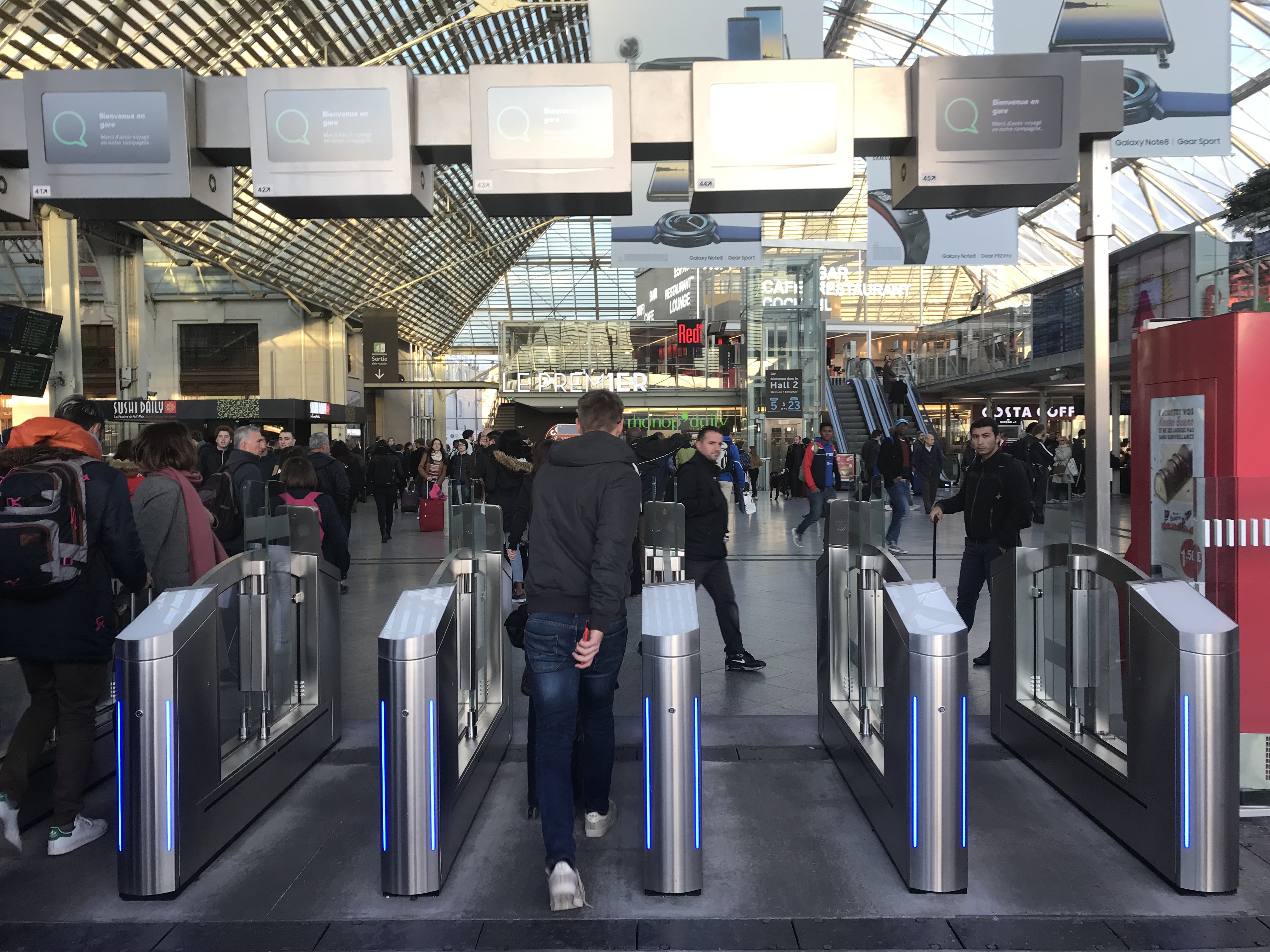 Eine Gruppe von Menschen geht durch einen belebten Bahnhof mit Metalldetektoren, Textanzeigen, Säulen, Rolltreppen und Drehkreuzen unter einer sichtbaren Decke.