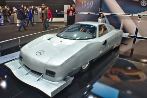 Ein silberner Mercedes-Benz 300 SL Roadster wird auf dem Frankfurt Motor Show präsentiert, umgeben von Menschen in verschiedenen farbigen Kleidern, mit Schildern und anderen Gegenständen im Hintergrund.