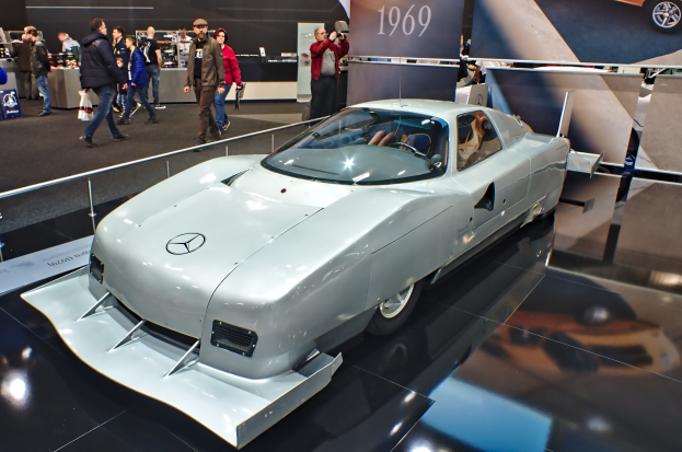 Ein silberner Mercedes-Benz 300 SL Roadster wird auf dem Frankfurt Motor Show präsentiert, umgeben von Menschen in verschiedenen farbigen Kleidern, mit Schildern und anderen Gegenständen im Hintergrund.