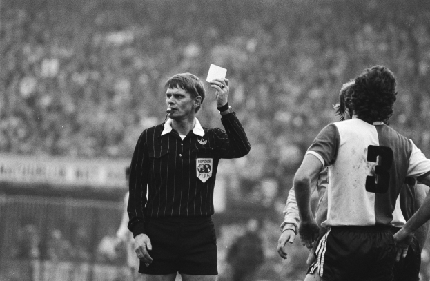 Ein Schiedsrichter in Schwarz-Weiß-Haltung hält eine rote Karte vor einer Menge von Menschen in Fußballtrikots, mit einem Zaun im Hintergrund.