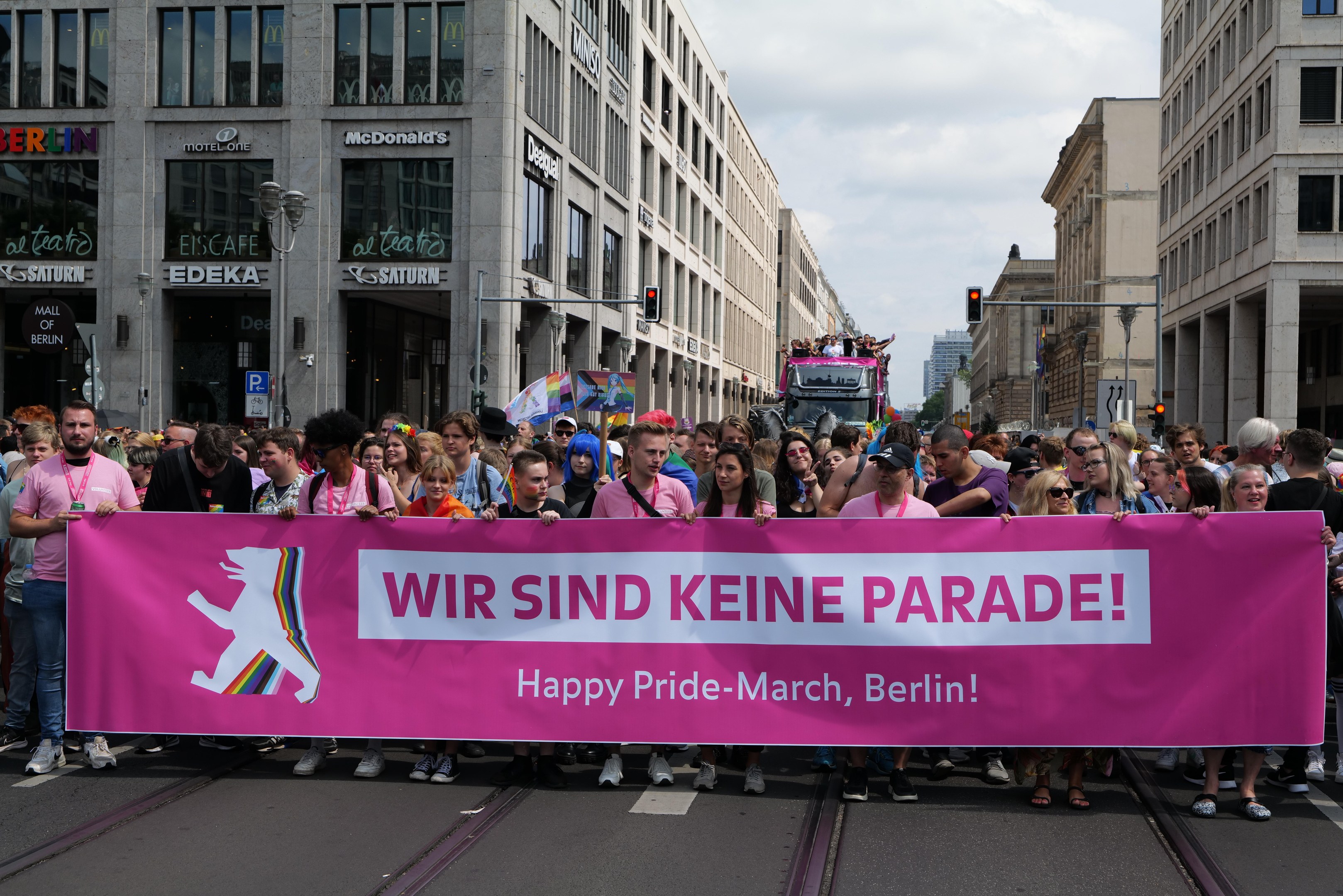 Eine Gruppe von Menschen marschiert auf einer Berliner Straße mit einem pinken Banner mit der Aufschrift "Happy Pride March", mit Gebäuden, Laternenmästen und Verkehrsampeln an der Straße entlang unter einem bewölktem Himmel.